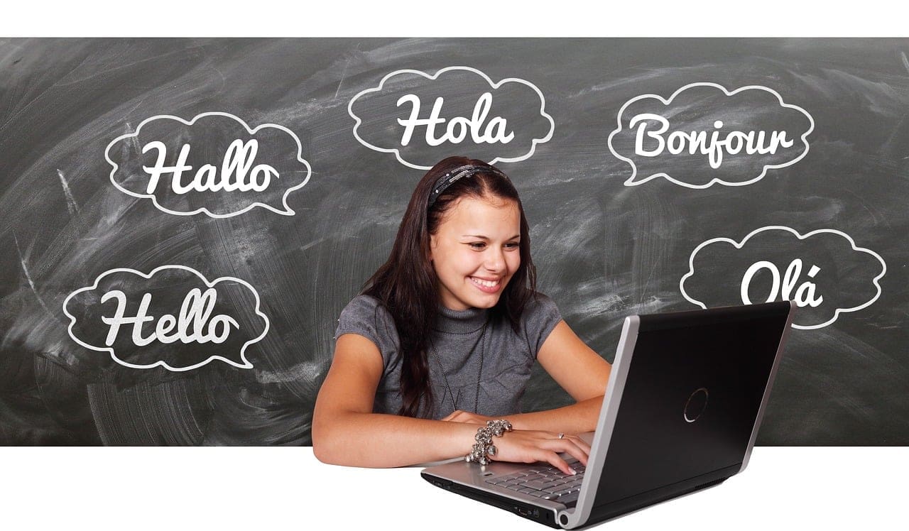 A woman looking at a laptop with a chalkboard featuring the word "hello" in several languages behind her.